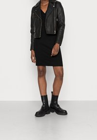 Giubbotto da biker in pelle nera con hardware argentato, indossato sopra un vestito nero a coste. Stivaletti neri con suola spessa completano l'outfit.