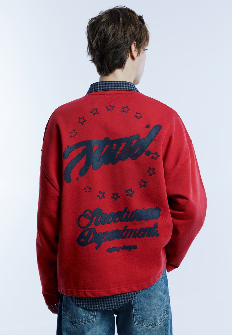 Rode oversized sweatshirt met zwarte graphics, waaronder tekst en sterren op de achterkant. Geribbelde manchetten, losse pasvorm. Gedragen over een geruit overhemd.