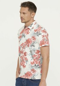 Polo à manches courtes en crème avec un motif floral rouge et vert. Il présente un col classique et une patte de boutonnage, fabriqué en tissu léger.