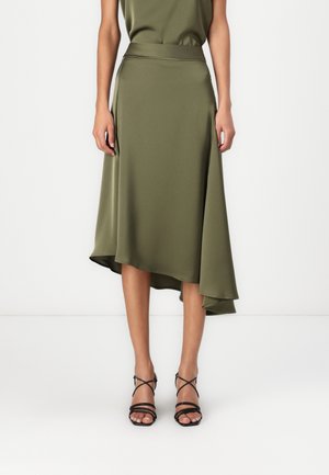 Olive green asymmetrical midi skirt with smooth fabric, worn with black strappy high-heeled sandals against a white background.