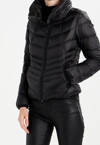 Mustapuffatakki, jossa on vetskarilla varustettu etuosa, korkea kaulus ja vaakasuuntainen tikkauskuvio. Sisältää sivutaskut ja elastiset hihnat.