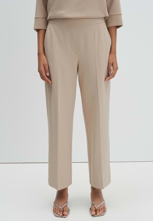 Vrouw die beige, recht gesneden, op maat gemaakte broek draagt en zilverkleurige sandalen met slangenhuidpatroon, open teen en hak, staand tegen een eenvoudige achtergrond.