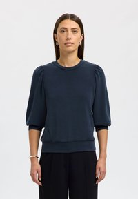 Granatowa bluza z okrągłym dekoltem i bufkowatymi rękawami, wykonana z miękkiego materiału. Posiada dopasowany dół i prosty design, noszona z czarnymi spodniami.