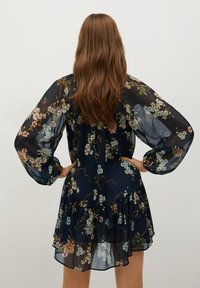 Nainen, jolla on pitkä ruskea hius, pukeutuneena tummansiniseen kukalliseen läpikuultavaan mekkoon, jossa on pitkät puffihihat. Hän seisoo kädet lanteilla ja selkä kameran suuntaan.