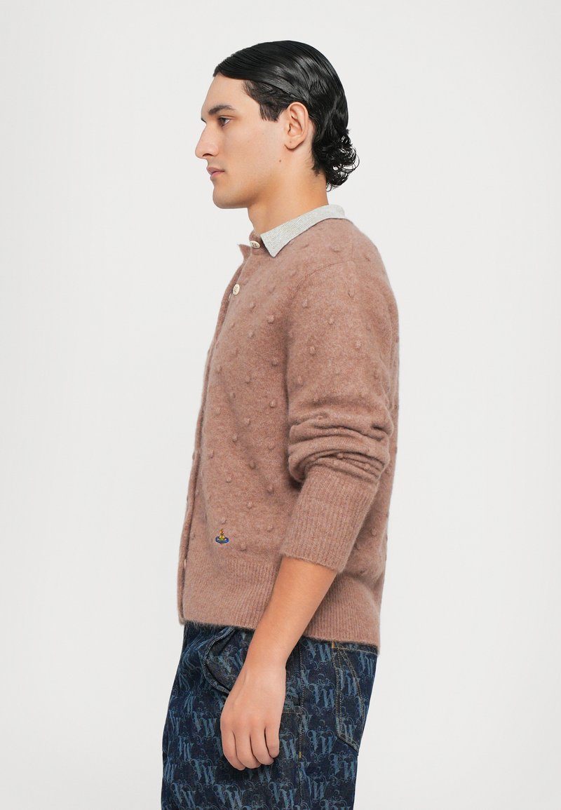 Bruine wollen cardigan met gestructureerde bobbels, voorzien van een knoopsluiting en geribde manchetten, gedragen over een lichtblauw hemd met kraag en met een patroon bedekte broek.