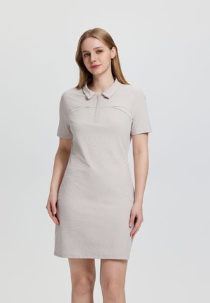 Młoda kobieta w beżowej sukience polo z krótkim rękawem i subtelnym wzorem zawijasa, stojąca ze swobodnie opuszczonymi ramionami na jednolitym jasnoszarym tle.