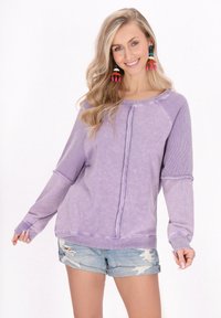Pulover lung cu mânecă lungă, de culoare mov, cu accente zimțate și margini destramate, asortat cu pantaloni scurți din denim uzat. Material moale cu o textură spălată.