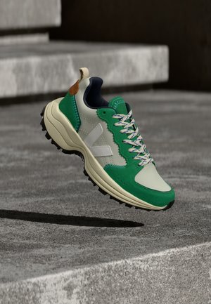 Sapatilha atlética verde e creme com acabamento texturizado, detalhe de logótipo branco, atacadores multicoloridos, sola de borracha e um destaque castanho no calcanhar.