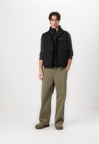 Black puffer vest over a dark green sweater, paired with olive green pants and brown boots. The vest features a high collar and snap closures.