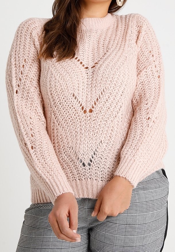 Pull en maille rose clair avec une coupe ample, un motif texturé et des détails en points ajourés, porté avec un pantalon à carreaux.