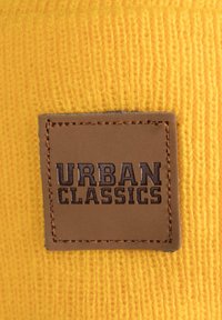 Material tricotat galben cu o textură canelată, având un etichetă dreptunghiulară din piele brună, debossată cu "URBAN CLASSICS."