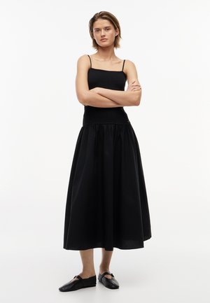 Młoda kobieta stoi ze skrzyżowanymi ramionami, ubrana w czarny top bez rękawów, długą czarną spódnicę i czarne płaskie buty na białym tle.