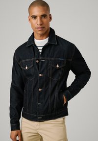 Chaqueta de mezclilla oscura con un diseño clásico, cierres de botones, bolsillos en el pecho y costuras amarillas en contraste, con un cuello estructurado.