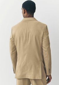 Blazer beige su misura con un unico spacchetto sul retro, texture liscia, rever a scollatura e accenti di bottoni in metallo sulle maniche.