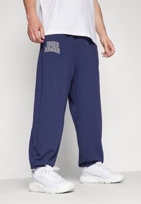 Navy blue sweatpants with elastic waistband, embroidered "UNDER ARMOUR" logo, relaxed fit, and a soft, textured fabric. Paired with white sneakers.