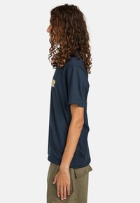 Marineblå bomulds t-shirt med korte ærmer, med et grafisk design på forsiden. Båret med olivengrønne cargo bukser, vist fra siden.