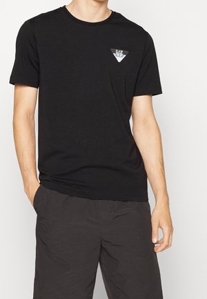 Homme portant un t-shirt noir à manches courtes avec un petit logo EA7 sur le côté gauche de la poitrine et un pantalon foncé, debout les mains partiellement dans les poches.