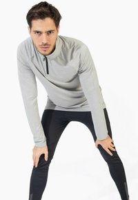 Camisa gris de manga larga con cremallera y textura acanalada, combinada con pantalones negros ajustados que presentan un diseño sutil. Modelo en una pose de estiramiento.