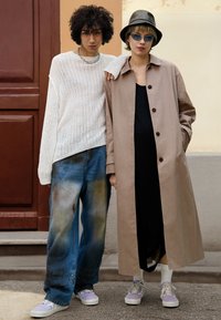 Deux jeunes posent côte à côte ; l'un porte un pull blanc et un jean large, l'autre un trench beige, une robe noire et un bob.
