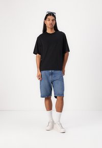 Svart kortermet t-skjorte med preget logo, kombinert med blå denimshorts. Modellen har på seg hvite sneakers og knehøye sokker.