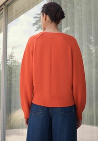 Oranger gestrickter Pullover mit Rundhalsausschnitt, Raglanärmeln und ribbeligem Saum; kombiniert mit blauen Denim-Hose im weiten Schnitt.