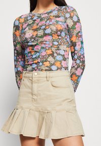 Blusa de manga larga con estampado floral colorido sobre un fondo oscuro, combinada con una falda acampanada beige con dobladillo con volantes y bolsillos frontales.