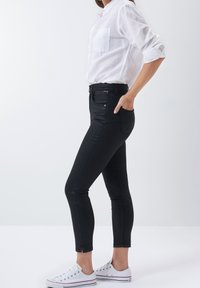 Black skinny jeans with a high waist, featuring front pockets, a zipper fly, and slight ankle slits. Paired with a white button-up shirt.