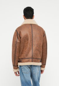 Uomo che indossa una giacca in shearling marrone con colletto e polsini in pelliccia crema, abbinata a jeans azzurri, voltato di spalle rispetto all'osservatore.