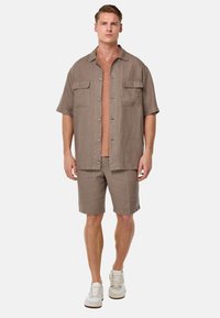 Camicia leggera in misto lino color taupe terroso con maniche corte e due tasche sul petto, abbinata a pantaloni corti coordinati e sneakers bianche.