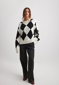 Schwarz-weißes Argyle-Muster-Sweatshirt mit geripptem Saum und weiten Ärmeln, kombiniert mit dunklen, engen Hosen und burgunderfarbenen Schuhen.