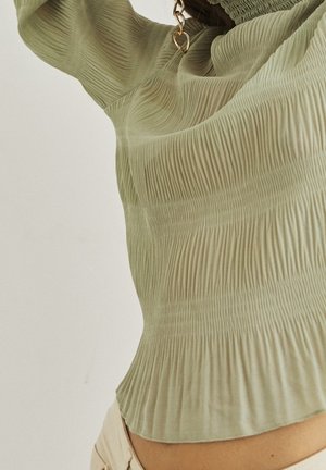 Lähikuva vaaleanvihreästä, teksturoidusta, laskostetusta pitkähihanaisesta paidasta, jota on yhdistetty beigeihin housuihin. Kuvassa näkyvät kankaan yksityiskohdat sekä istuvuus vyötäröllä ja käsivarressa.