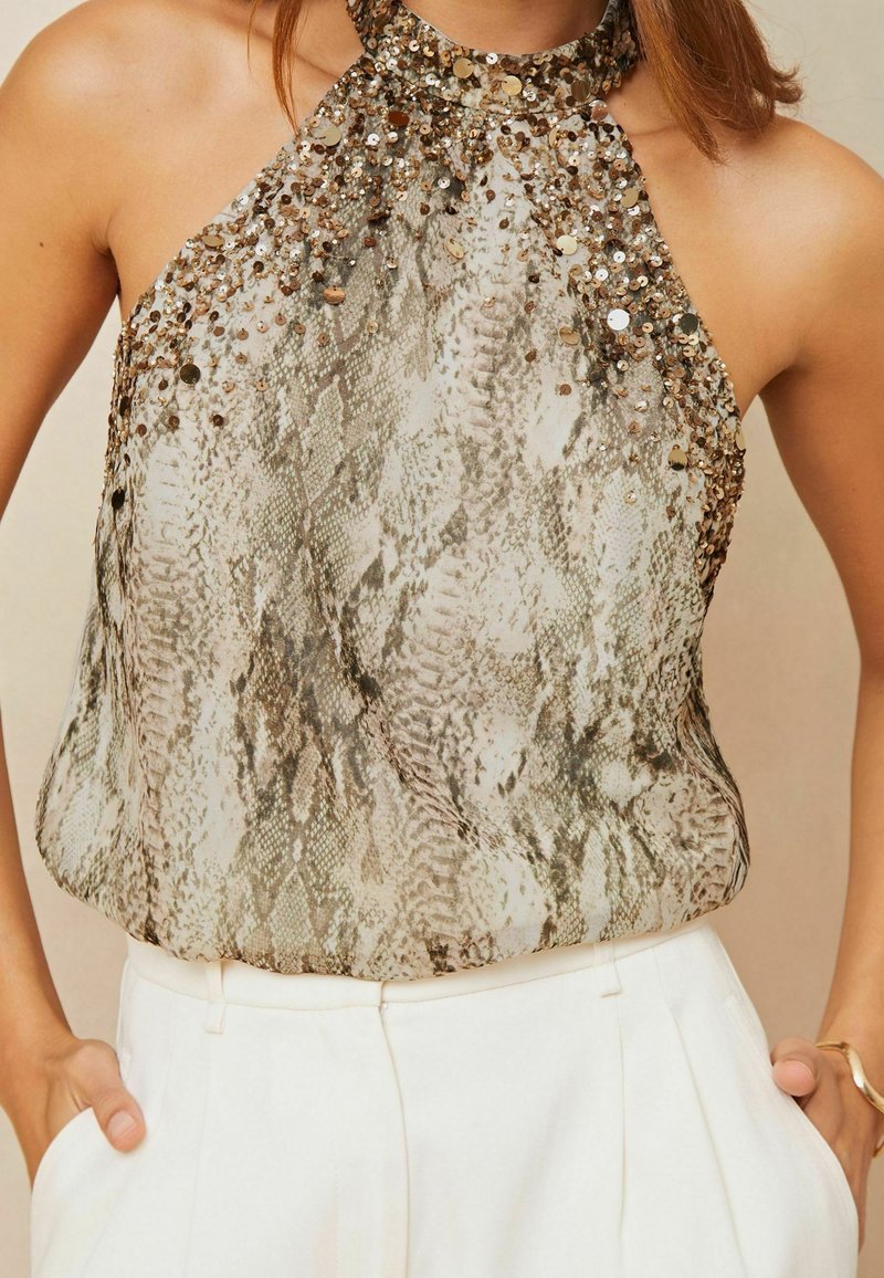 Top con cuello halter que presenta un patrón de serpiente, decorado con lentejuelas en tonos dorados y bronce. Tejido fluido y texturizado, combinado con pantalones de color claro.