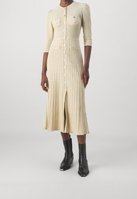Stickad klänning i ljusbeige med texturerat mönster, knappstängning fram, två bröstfickor och en medellång utsvängd kjol. Matchad med svarta stövlar.