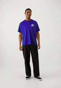 Vyras, vilkintis violetinį marškinėlį su grafika, juodas plačias kelnes, juodus sportinius batelius ir tonuotus akinius, stovi prieš vienodą baltą foną.