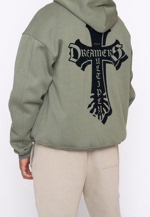 Osoba ubrana w oliwkowozieloną bluzę z kapturem z czarnym krzyżem i napisami "DREAMERS" oraz "MULTIPLY" na plecach, w połączeniu z beżowymi spodniami.