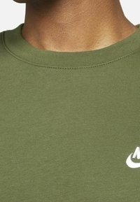 Närbild av en persons hals och övre bröst som bär en olivgrön rundhalsad sweatshirt med en delvis vit Nike-logga.