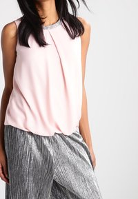 Blusa sin mangas rosa con pliegues y un escote acentuado con lentejuelas plateadas, combinada con pantalones anchos de pierna plisada de color plateado brillante.