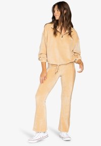 Tan corduroy lounge set met een gevoerde pullover met ritssluiting en elastische manchetten, gecombineerd met bijpassende flared broek en witte sneakers.