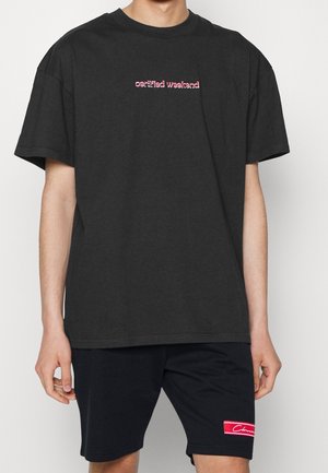 Osoba ubrana w czarny, oversize'owy t-shirt z napisem "certified weekend" oraz czarne spodenki z czerwoną, prostokątną naszywką z logo.