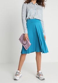 Camicia a fantasia blu chiaro, gonna midi plissettata turchese brillante, pochette lavanda con hardware circolare e sneaker bianche con accenti pastello.