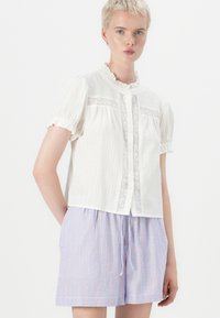 Hvid kortærmet bluse med blonderelaterede detaljer og knaplukning fortil. Sammen med lyseblå stribede shorts med røde accenter.