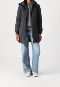 Abrigo de plumas negro largo con capucha, puños acanalados y cierre de cremallera. Combinado con un suéter gris y unos pantalones vaqueros de pierna ancha de color azul claro.