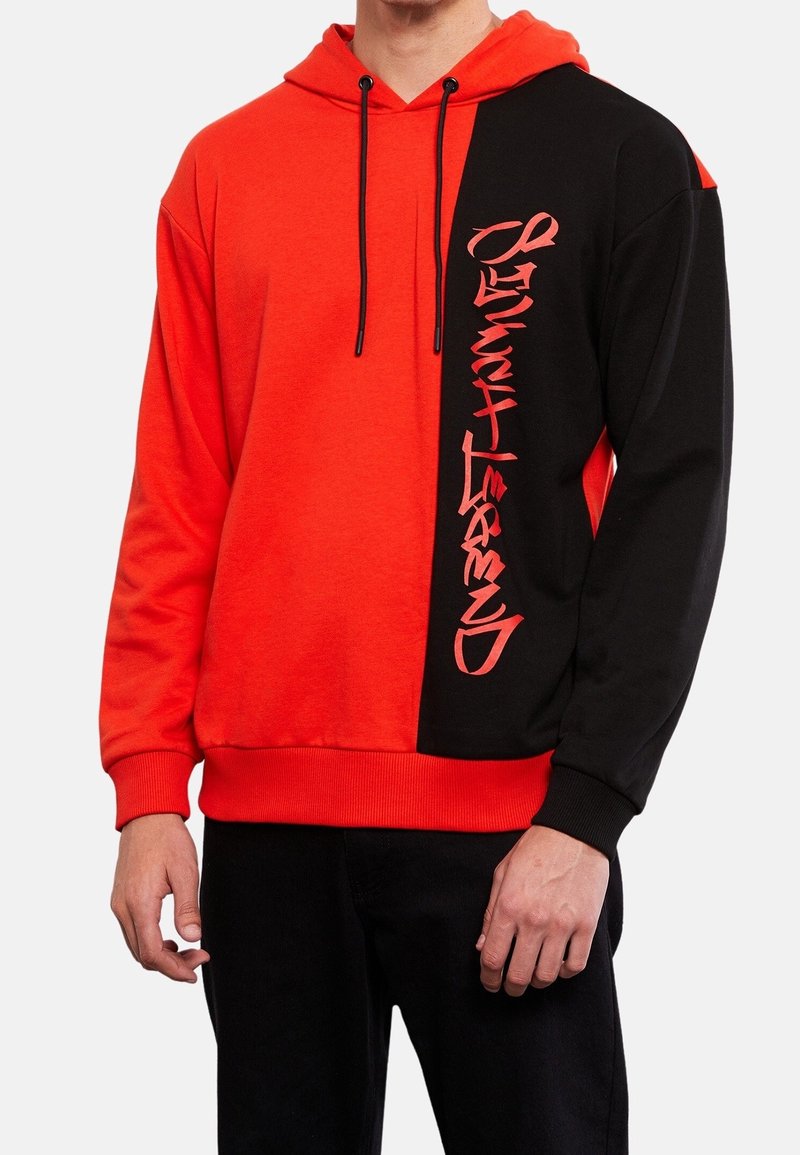 Homme portant un sweat à capuche rouge et noir avec un texte vertical de style graffiti sur le panneau noir, debout les bras détendus le long du corps.