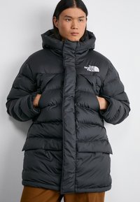 Giacca a vento nera con colletto alto, dotata di chiusura frontale con zip, due tasche anteriori e un logo sul lato sinistro del petto.