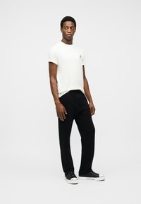 Άνδρας που στέκεται, φοράει λευκό T-shirt με κοντά μανίκια, μαύρο παντελόνι με κορδόνι στη μέση και μαύρα αθλητικά παπούτσια χαμηλού τύπου, κοιτάζοντας ελαφρώς προς τα δεξιά, σε απλό φωτεινό φόντο.