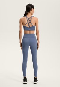 Kék sportmelltartó, keresztpántos dizájnnal és magas derekú, hozzá illő leggings, mindkettő rugalmas anyagból készült, fekete sportcipővel párosítva.