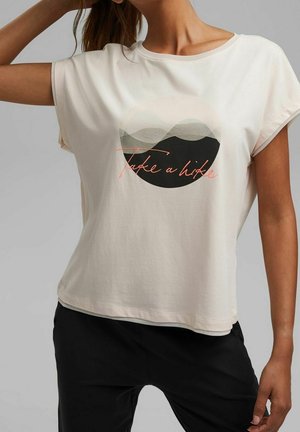Femme portant un t-shirt blanc avec un graphique circulaire de montagne et le texte "Take a hike" en écriture cursive.