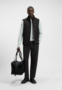 Svart quiltad väst över en ljusblå långärmad skjorta, ihop med svarta byxor och sneakers. En svart väska med en böjd dragkedja hålls.