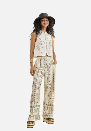 Mujer con un sombrero negro de ala ancha, chaleco sin mangas de color crema bordado, pantalones de pierna ancha con estampado, sandalias de plataforma y pulsera dorada.