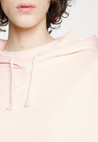Sudadera rosa claro hecha de tejido suave, con capucha ajustable, logo de Nike en blanco y detalle texturizado en el dobladillo y las mangas.